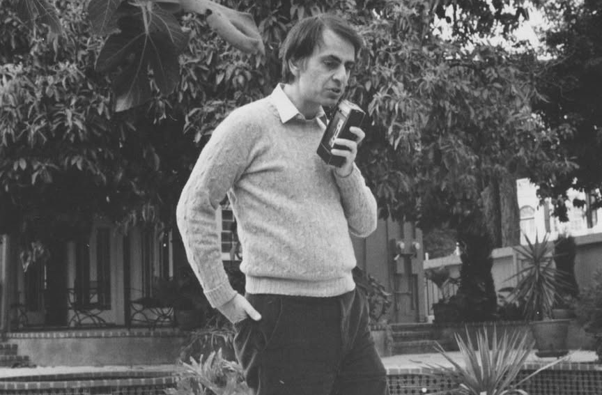 Fotografia em preto e branco de Carl Sagan ao ar livre, segurando um livro aberto perto do rosto.